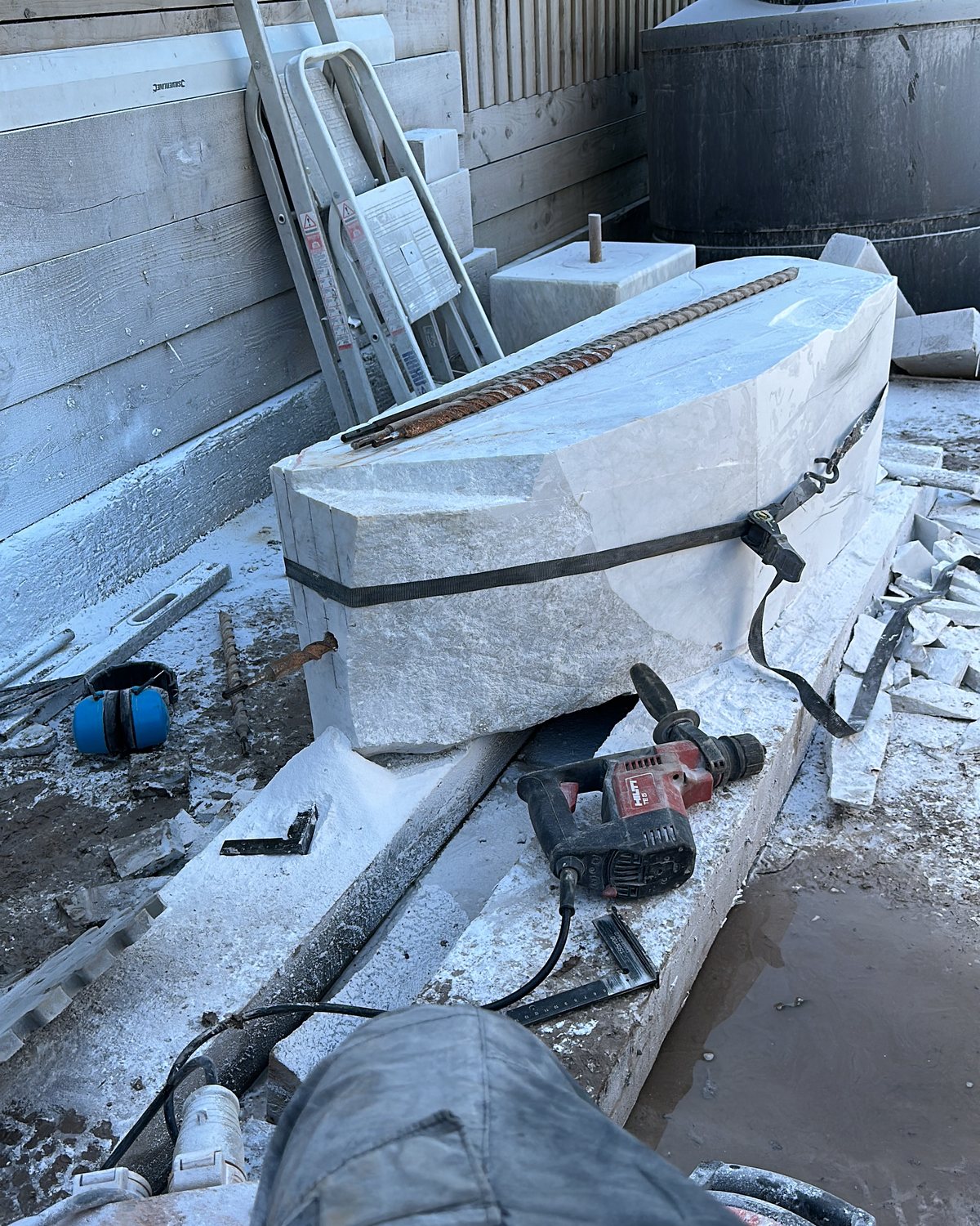 A long block of pale marble in the yard with a long drill bit resting on top, a hammer drill and ear defenders beside it