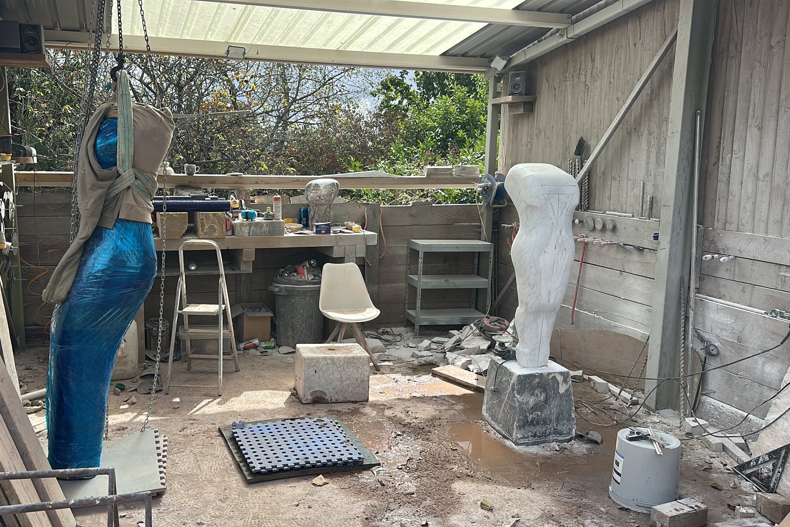 Inside the workshop: a wrapped piece hanging from the chain hoist, a white figure marked up in pencil on a block, and stone dust across the floor